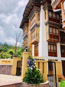 A traditional Bhutanese-style building with ornate wooden windows, colorful details, and flowered plants at the entrance. A sign reads OVERLAND. Cloudy sky and green hills are visible in the background.