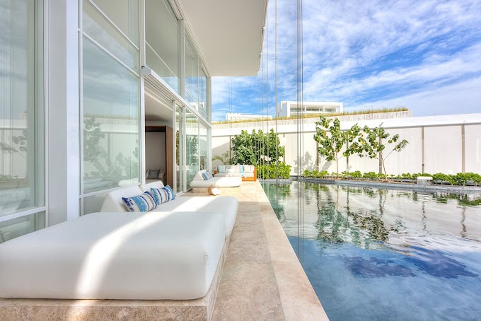 A modern poolside lounge area with white cushioned daybeds, glass walls, and clear blue water, surrounded by greenery and bright sky with clouds.