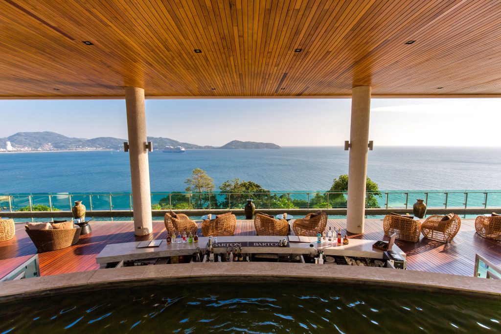 Outdoor bar with wicker chairs and tables on a wooden deck, overlooking a blue ocean and distant islands, framed by tall columns and a wooden ceiling. Glass railings provide an unobstructed view of the scenic coastline.