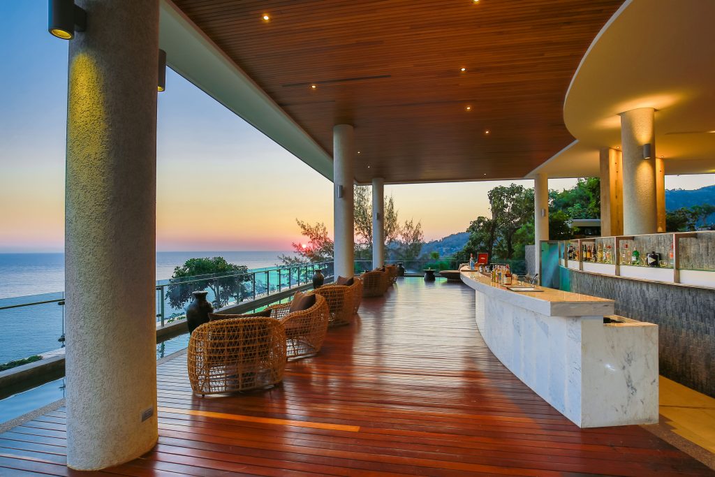 A modern, open-air lounge with wicker chairs and a marble bar overlooks the ocean at sunset. The wooden ceiling and floor create a warm ambiance, while glass railings offer an unobstructed view of the sea and sky.