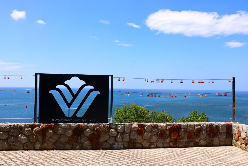 A stone terrace overlooks the ocean under a clear blue sky, with a Wyndham Grand Phuket sign and a fence decorated with colorful love locks.