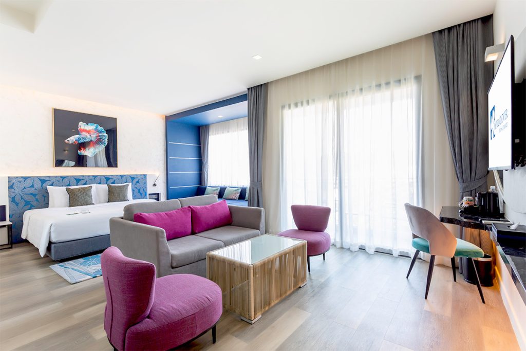 A modern hotel room with a large bed, colorful seating area with purple chairs and a pink sofa, a glass coffee table, a work desk, and large windows with sheer curtains letting in natural light.