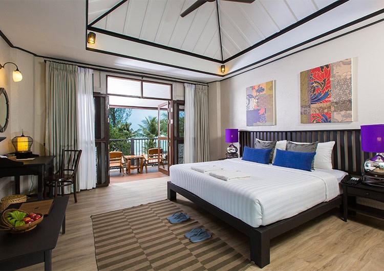 A spacious hotel room with a large bed, colorful pillows, two bedside tables with purple lamps, wall art above the bed, a desk with a mirror, and a balcony overlooking palm trees and the ocean.
