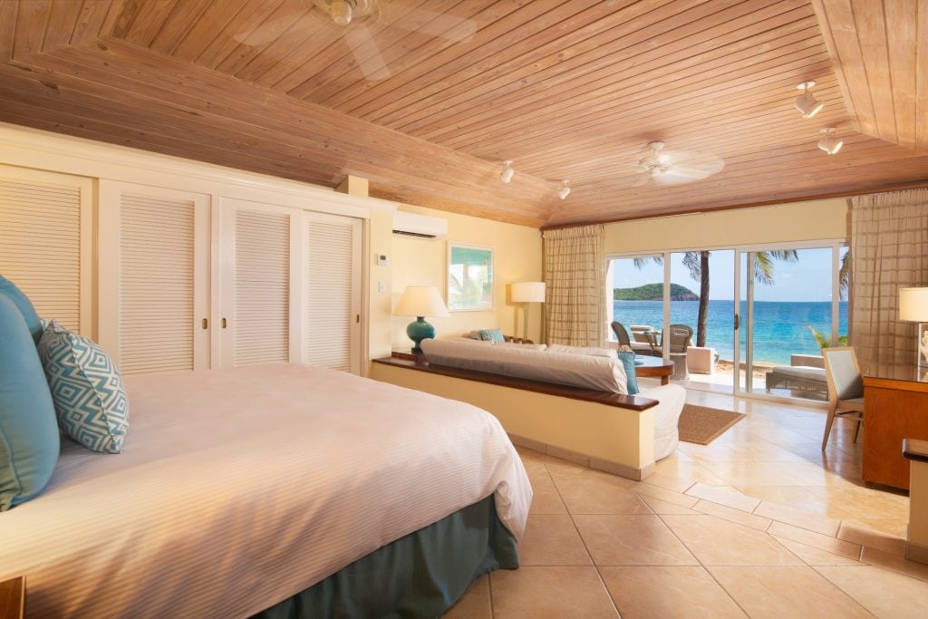 A spacious, sunlit hotel room with a large bed, sofa, and desk. Sliding glass doors open to a patio with tropical views of palm trees, a sandy beach, and turquoise ocean water. Light wood accents create a relaxing atmosphere.