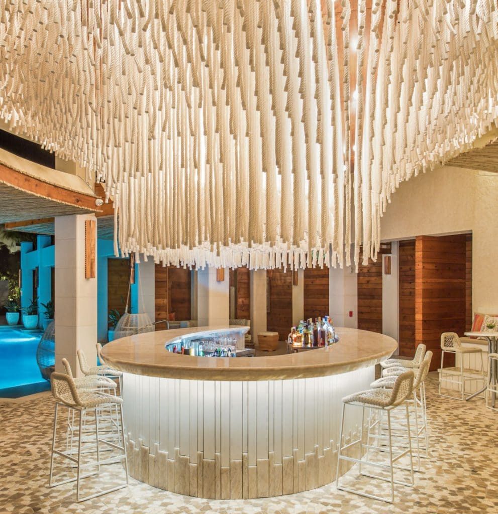 A modern, circular bar with a glowing counter and three barstools is beneath a dramatic ceiling display of hanging white ropes. Bottles and glasses sit on the bar, and a pool area is visible in the background.