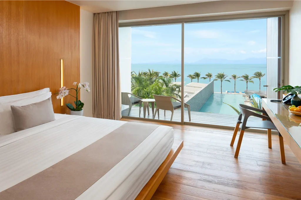 A modern hotel room with a neatly made bed, wooden floors, a desk with fruit, and a private balcony overlooking a pool, palm trees, and the ocean under a clear sky.