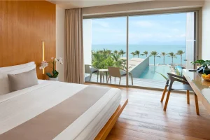 A modern hotel room with a neatly made bed, wooden floors, a desk with fruit, and a private balcony overlooking a pool, palm trees, and the ocean under a clear sky.