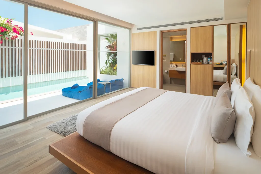 Modern hotel room with a large bed, neutral bedding, and wooden accents. Floor-to-ceiling glass doors open to a private patio with a pool, blue loungers, and vibrant flowers. Bathroom and minibar area visible.