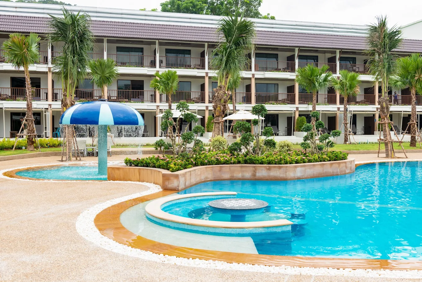 A resort-style swimming pool with a blue mushroom-shaped fountain, palm trees, and a two-story hotel building with balconies in the background. The pool area includes a small circular seating section.