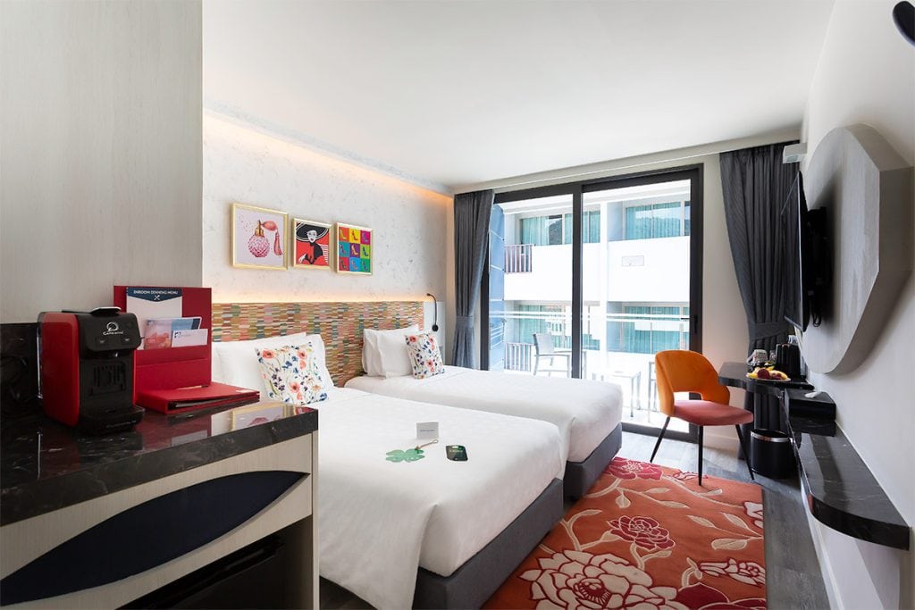 Modern hotel room with two white twin beds, colorful pillows, wall art, a red coffee machine, a small table with a chair, and large windows with a balcony view.