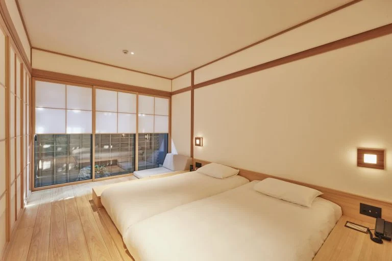 A minimalist Japanese-style bedroom with two low twin beds, light wood flooring, white walls with wood trim, large shoji screen windows, and a small sitting area near the window.