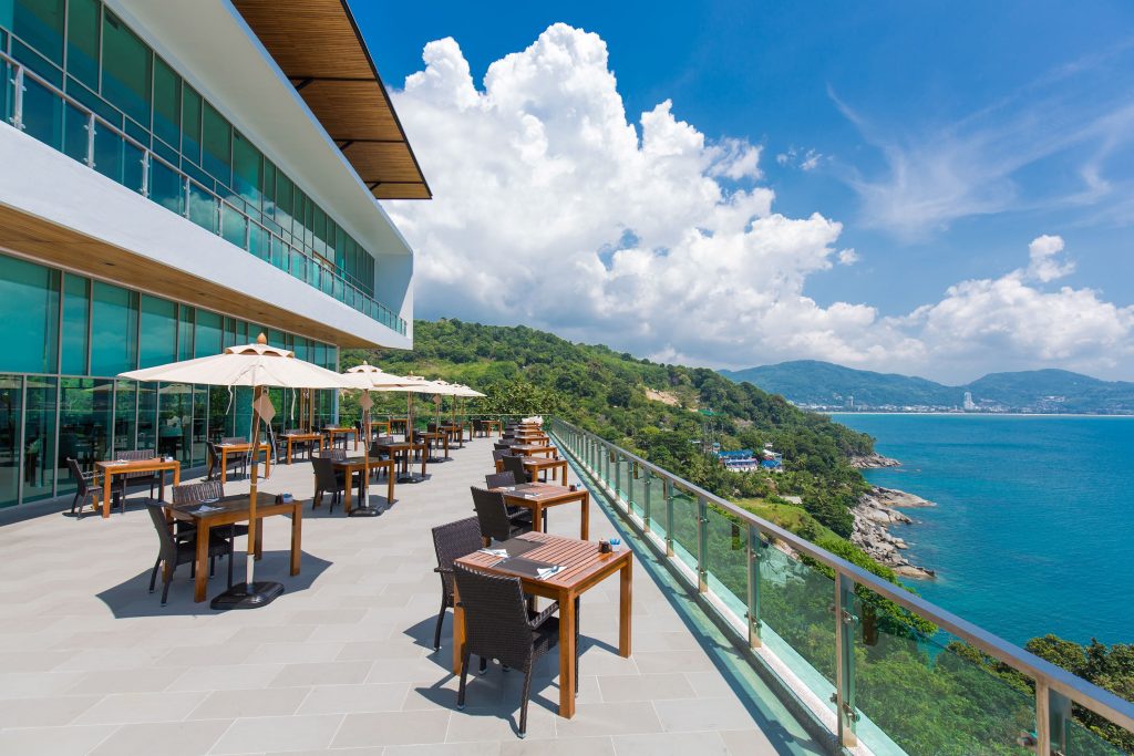 An outdoor restaurant terrace with tables and umbrellas overlooks a scenic coastline, lush green hills, and blue ocean under a bright sky with scattered clouds.