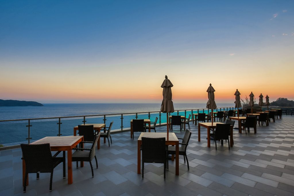 Outdoor restaurant terrace with empty tables and chairs overlooks a calm ocean at sunset. Closed umbrellas line the railing, and the sky is a gradient of blue to orange. The scene is peaceful and inviting.