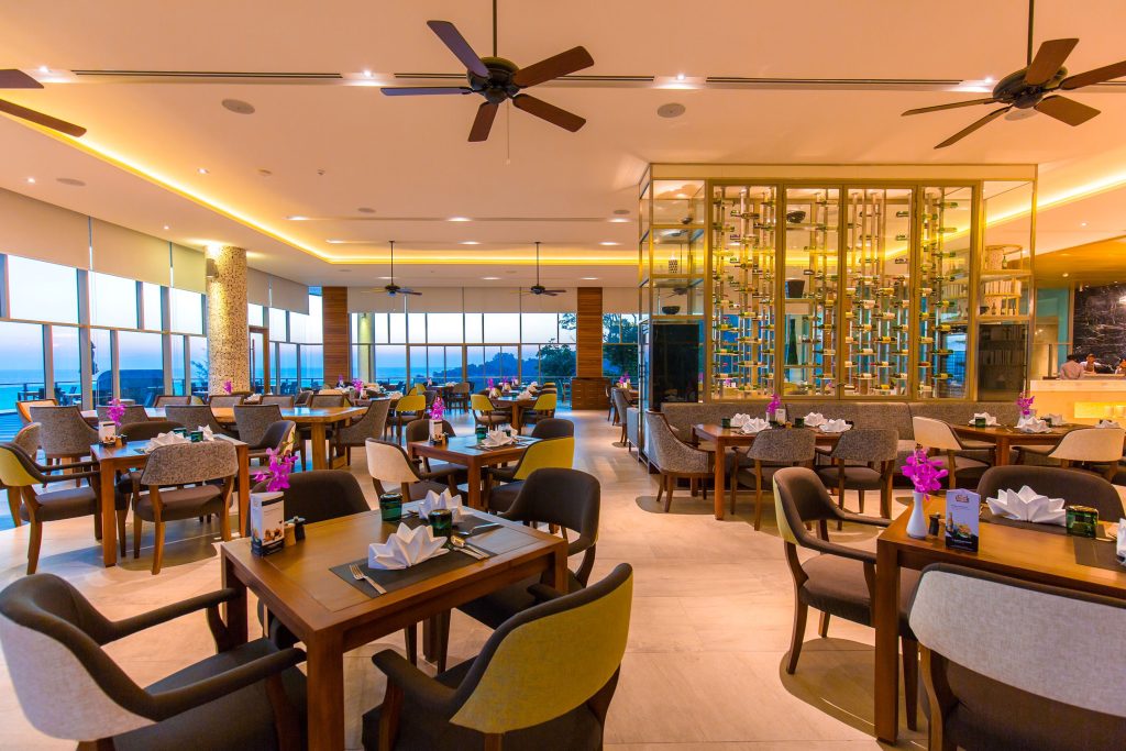 A spacious, modern restaurant with wooden tables and cushioned chairs, decorated with purple flowers. Large windows show an outdoor view, and ceiling fans hang above. Soft, warm lighting creates a cozy atmosphere.
