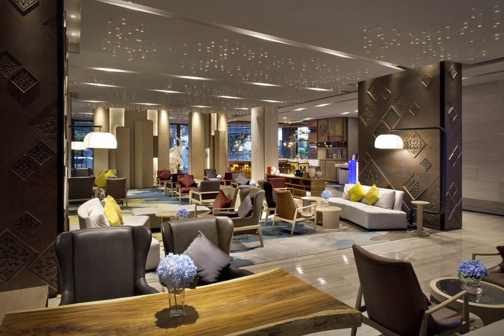 Modern hotel lobby with elegant seating areas, decorative lighting, and contemporary furniture. Sofas and chairs are arranged around tables, with cushions and flowers adding color to the stylish, open space.