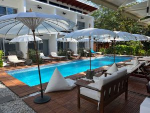 A serene outdoor pool area with white lounge chairs, wooden deck, and large white umbrellas. Modern white building with balconies is in the background, surrounded by greenery and sunlight.
