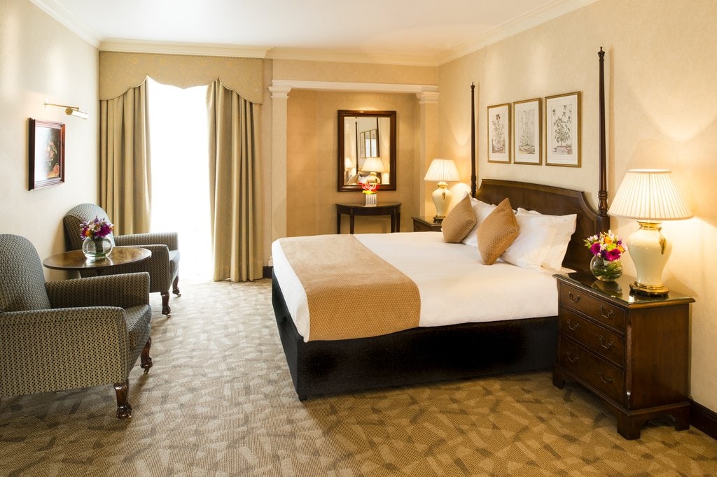 A spacious hotel room with a large bed, tan and white bedding, two nightstands with lamps, a table with two armchairs, framed artwork on the wall, and a window with beige curtains letting in natural light.