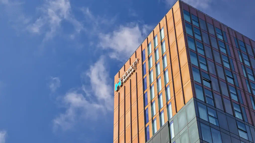 A modern high-rise building with an orange and glass exterior stands against a blue sky with scattered clouds. The TransUnion logo is visible near the top corner of the building.