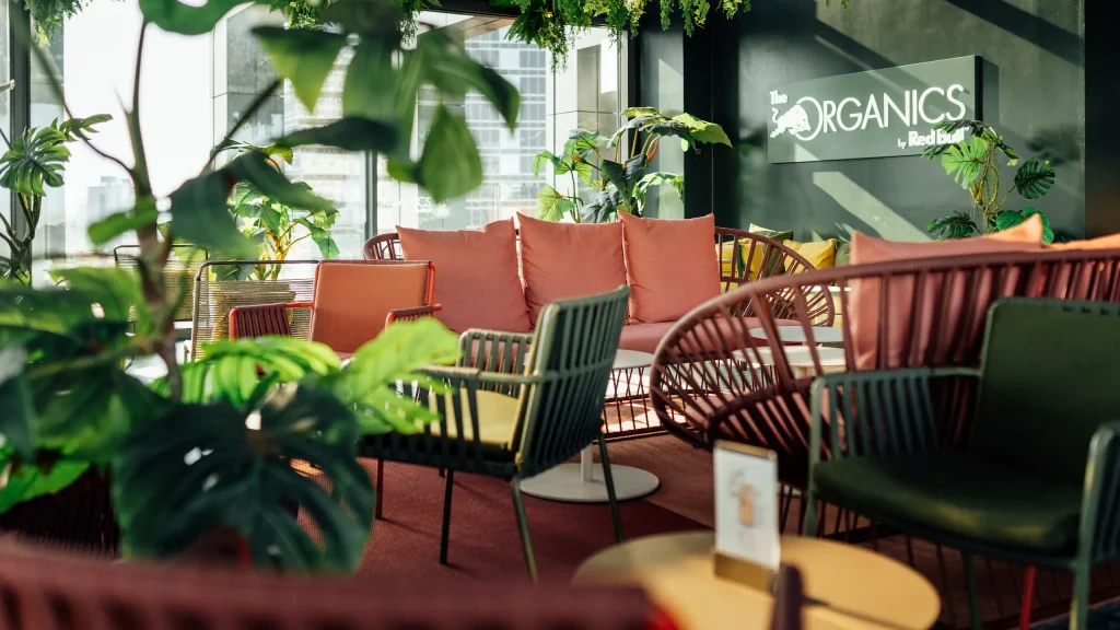 A modern lounge area with colorful chairs, sofas with red cushions, indoor plants, and sunlight streaming in. A sign on a dark wall reads “ORGANICS by Red Bull.”.