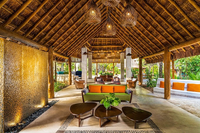 Open-air tropical lounge with a thatched roof, wicker lamps, green and orange cushioned sofas, modern wooden tables, a stone wall with a water feature, and lush greenery visible outside.