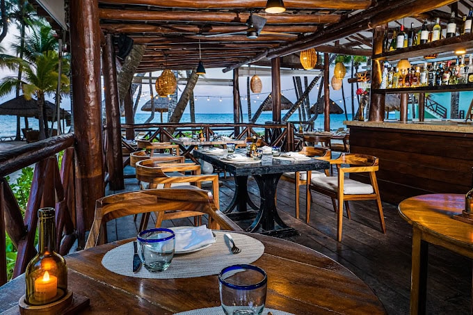 Open-air seaside restaurant with wooden tables and chairs, set for dinner. Hanging woven lamps create a warm ambiance. Bar area visible on the right. Palm trees, thatched umbrellas, and ocean view in the background.