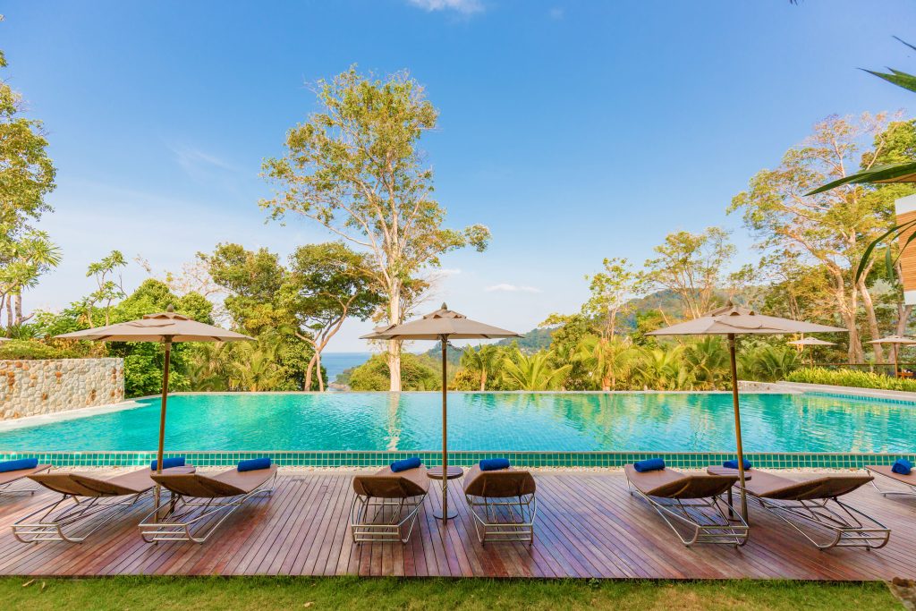 Lounge chairs and umbrellas line a wooden deck beside a large, clear swimming pool, surrounded by trees and lush greenery, with a scenic view of hills and blue sky in the background.