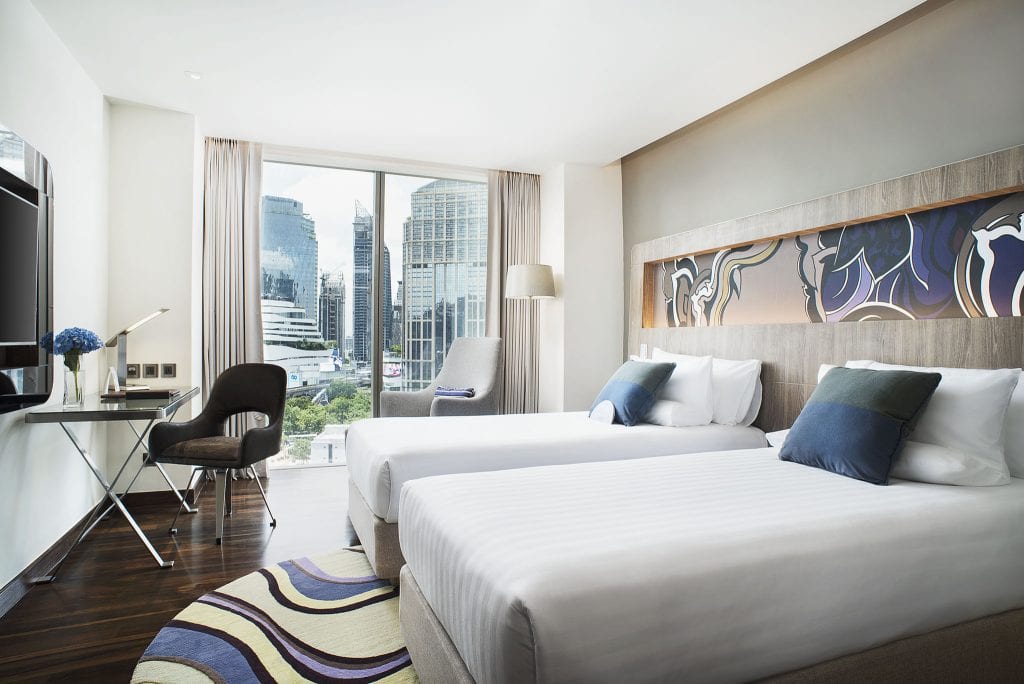Modern hotel room with two twin beds, a desk and chair, an armchair by tall windows, city buildings visible outside, and stylish decor in neutral tones with blue accents.