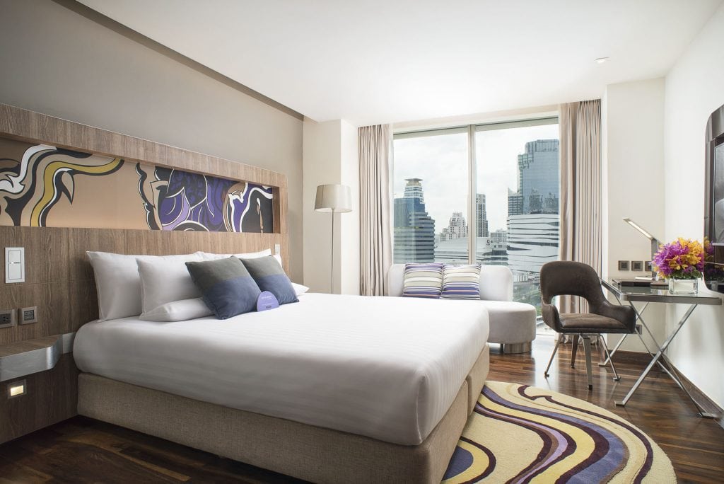 A modern hotel room with a large bed, colorful pillows, abstract wall art, a white sofa, a desk with flowers, and a window showing a city skyline with tall buildings.
