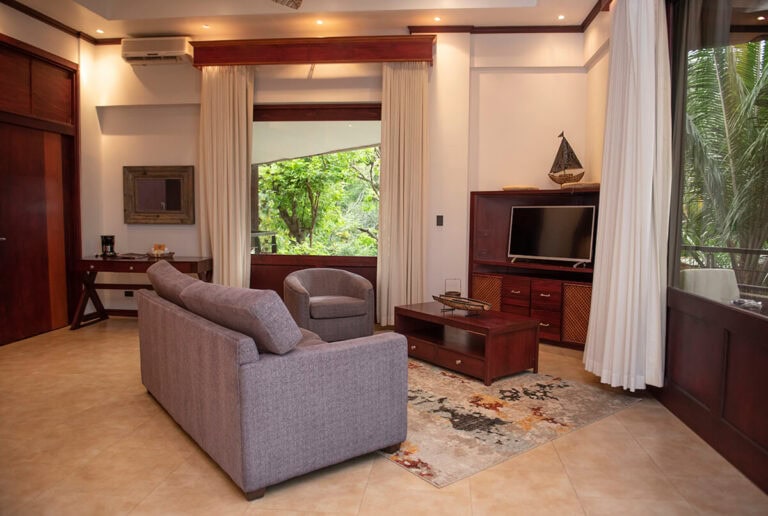 A cozy living room with a gray sofa, armchair, TV on a wooden stand, desk, and large windows with cream curtains, overlooking greenery outside. A decorative rug and model sailboat add homey touches.