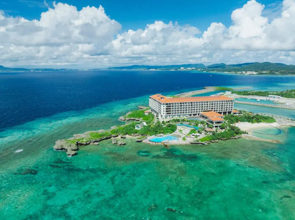 A large hotel with a red roof sits on a small, lush peninsula surrounded by clear turquoise water and coral reefs, with a swimming pool nearby and distant green hills under a partly cloudy sky.