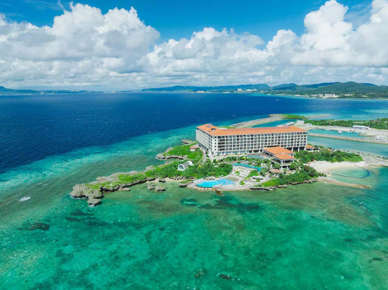 A large hotel with a red roof sits on a small, lush peninsula surrounded by clear turquoise water and coral reefs, with a swimming pool nearby and distant green hills under a partly cloudy sky.