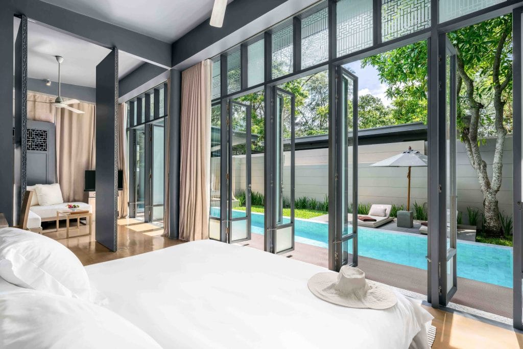 A modern bedroom with large glass doors opens to a private patio with a swimming pool, sun loungers, and an umbrella. Sunlight fills the room, highlighting a bed with white linens and a sunhat on it.