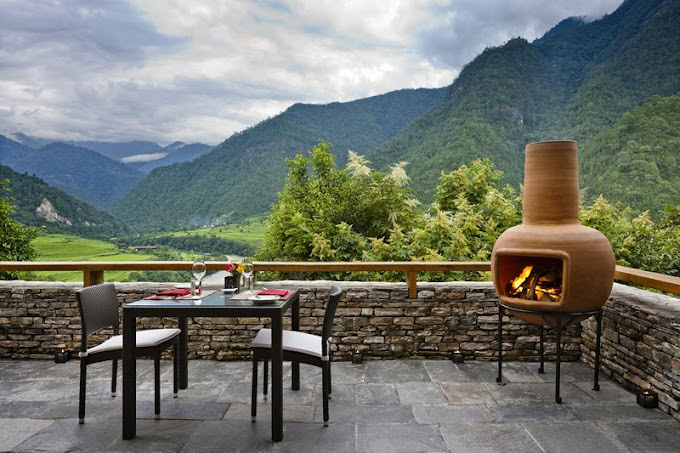 A stone patio with a table set for two and a lit outdoor fireplace overlooks lush green hills and mountains under a cloudy sky.