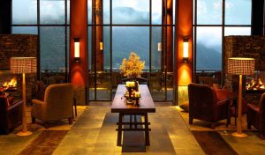 A cozy lounge with armchairs and a central wooden table adorned with candles and flowers, illuminated by warm lighting. Large windows reveal a foggy mountain view outside.