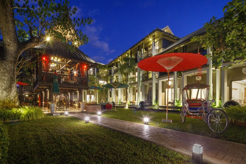 Courtyard of a hotel at dusk with illuminated walkways, a large red umbrella, a decorative rickshaw, lush green lawn, trees, and multi-story buildings featuring balconies and warm lighting.