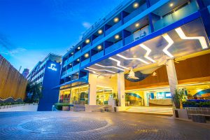 A modern hotel entrance at dusk with bright, stylish lighting, glass balconies, and illuminated signs. The building features clean lines, large windows, and a welcoming, well-lit driveway.