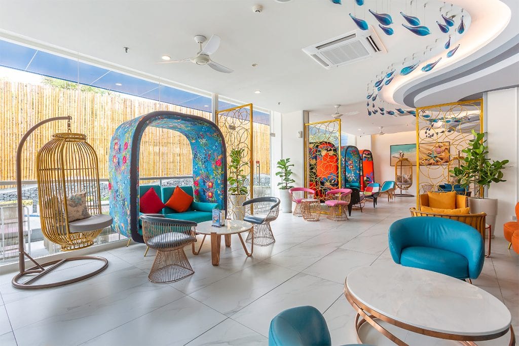 A bright, modern lounge with colorful chairs, hanging wicker seats, floral-patterned booths, marble tables, and large windows. Decorative peacock feather art adorns the walls and ceiling. Green plants add a fresh touch.