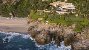 A luxurious villa sits on a lush, rocky cliff overlooking the ocean, with a sandy beach, sun loungers, palm trees, and waves crashing against the rocks below.