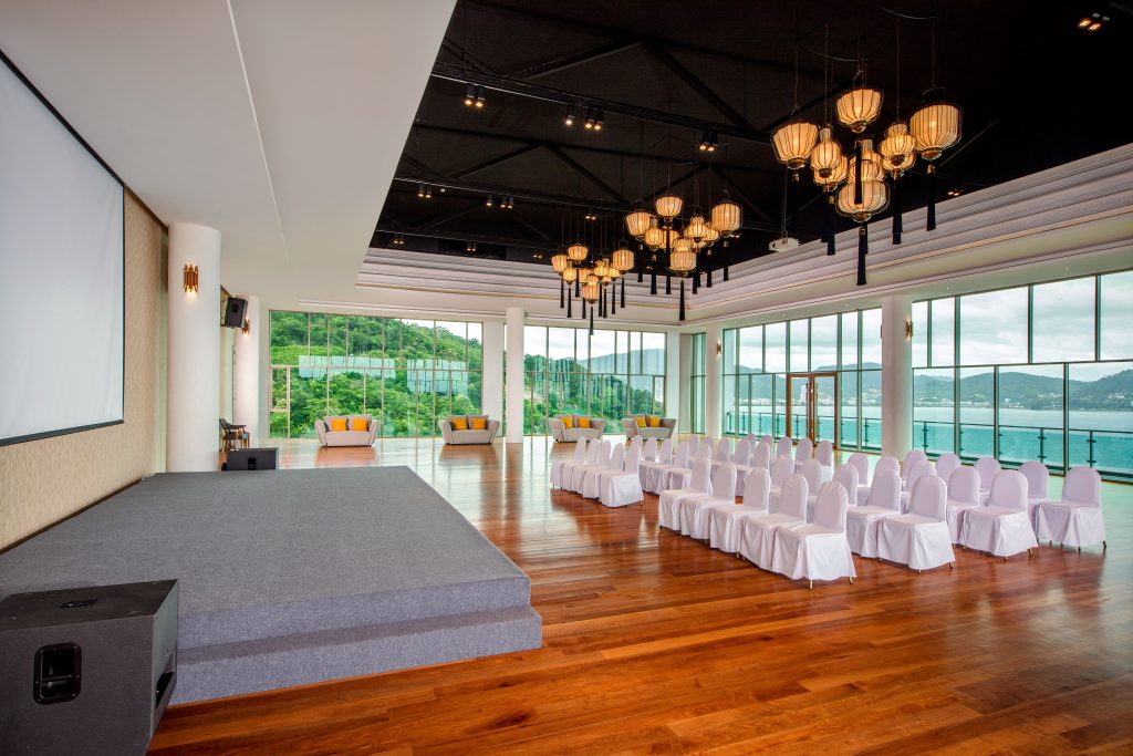 A spacious, modern event venue with floor-to-ceiling windows overlooking the ocean, a stage on the left, rows of white covered chairs, wooden floors, and decorative ceiling lights.