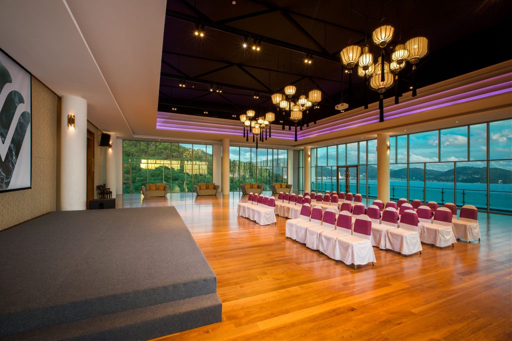 A modern event space with rows of purple and white chairs facing a stage, large windows with views of water and mountains, wooden floors, and elegant hanging lights.