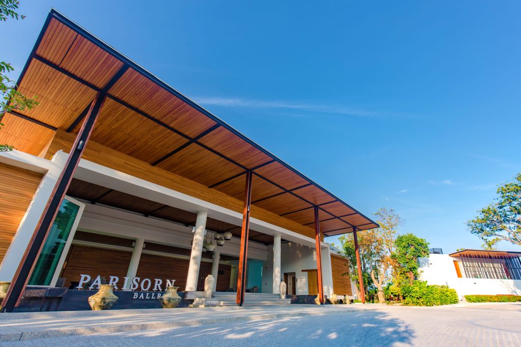 Modern building with a large wooden roof, tall pillars, and stone planters at the entrance. The sign reads Parisorn Ball. Clear blue sky and trees surround the area, creating a bright and welcoming atmosphere.