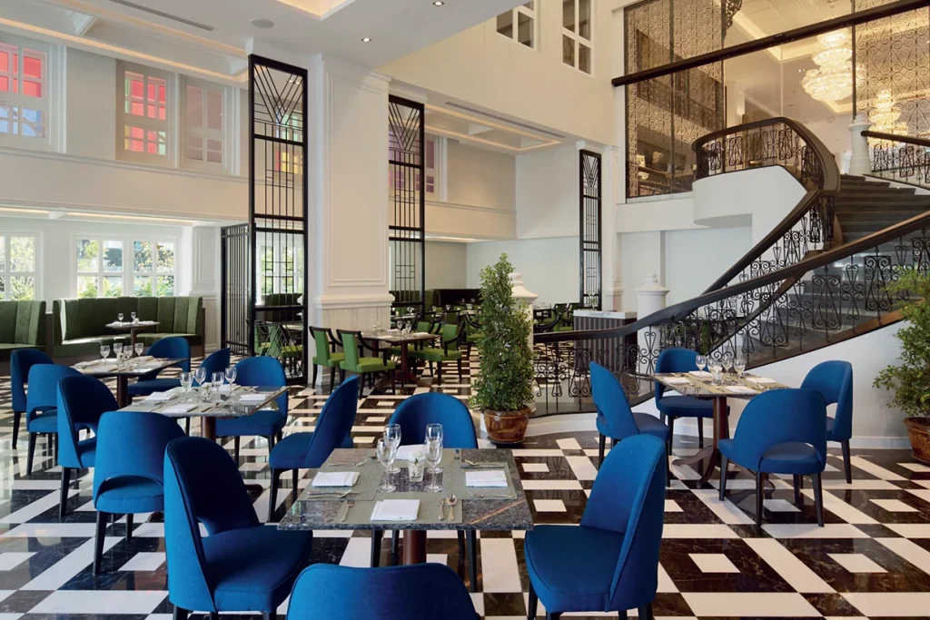 Elegant restaurant interior with black-and-white checkered floor, blue chairs around set tables, potted plants, and a grand staircase with ornate railing. Large windows and modern decor create a bright, stylish atmosphere.