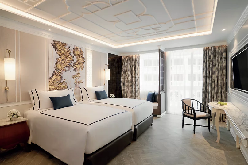 A modern hotel room with two twin beds, white bedding, blue accent pillows, a large map artwork on the wall, a desk with a chair, a flat-screen TV, and floor-to-ceiling windows with patterned curtains.