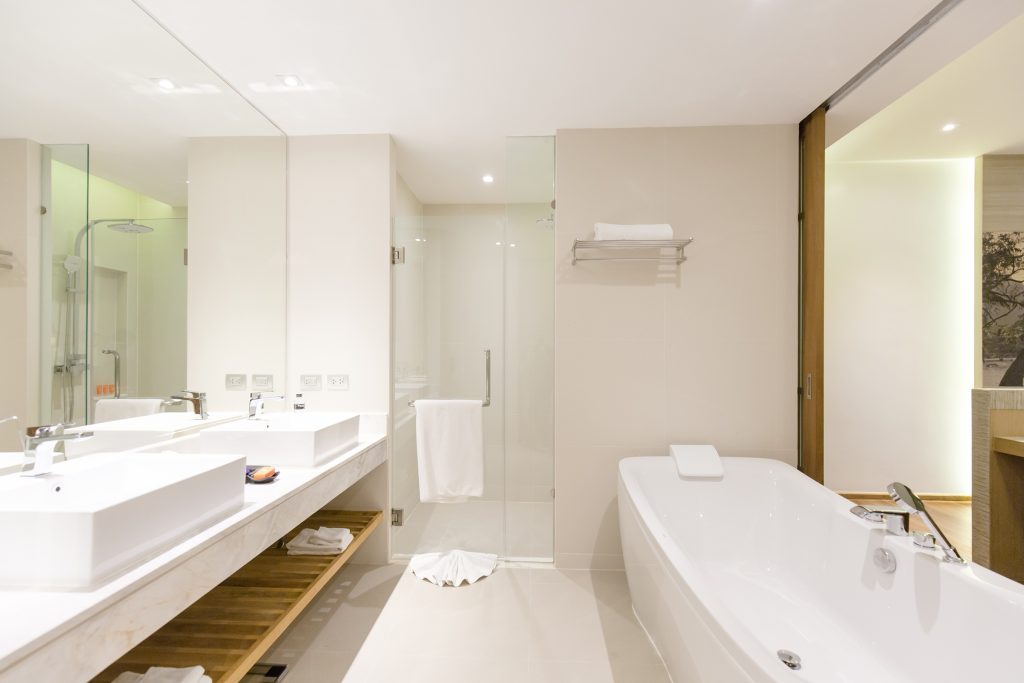 Modern bathroom with a large white bathtub, double sinks with mirrors, a walk-in glass shower, and neatly folded towels on a shelf. The space is bright with clean, minimalist decor.