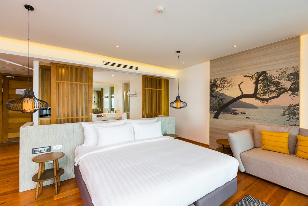 Modern hotel room with a large bed, white linens, wooden floors, a small sofa with yellow cushions, pendant lights, and a large wall art of a beach scene behind the bed. A bathroom area is visible in the background.