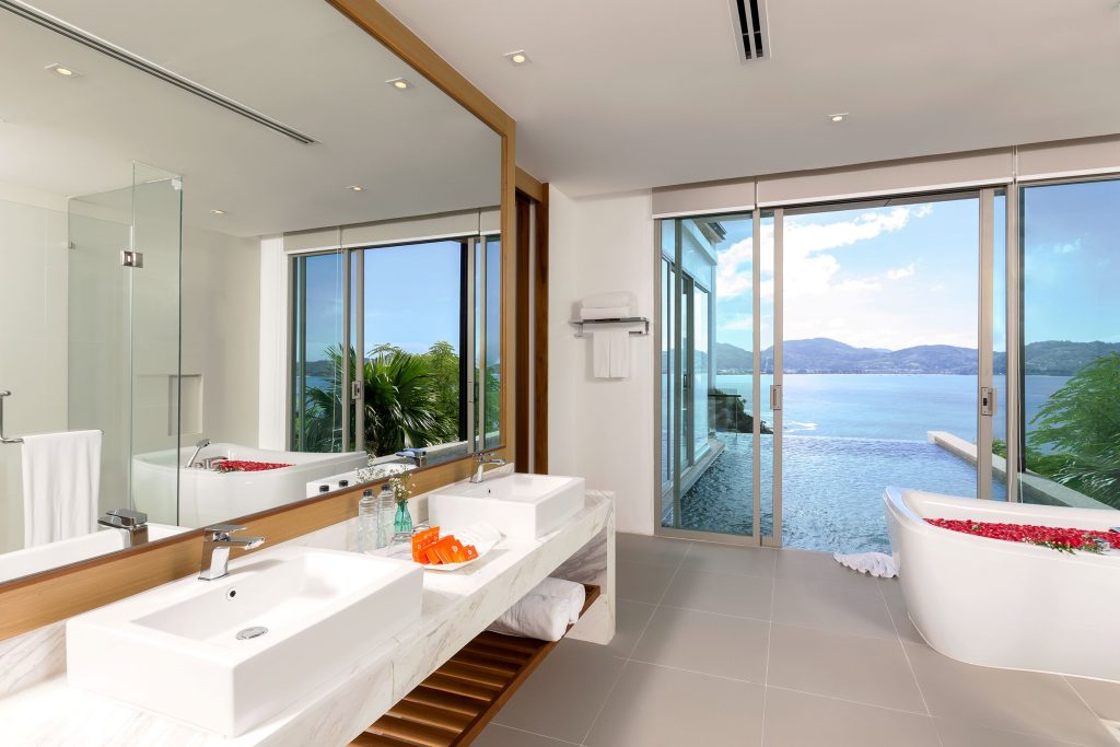 Modern bathroom with double sinks, large mirror, freestanding tub with rose petals, glass shower, floor-to-ceiling windows, and a view of an infinity pool overlooking a lake or sea with mountains in the background.