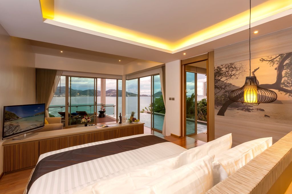 A modern hotel room with a large bed, TV, seating area, and floor-to-ceiling windows overlooking the ocean. The room has warm lighting, wood accents, and access to a private pool with scenic views.