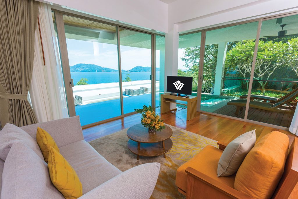 A bright, modern living room with a sofa, armchairs, and a coffee table overlooks a private pool and a scenic ocean view through large glass doors. A small TV sits near the window, and greenery is visible outside.
