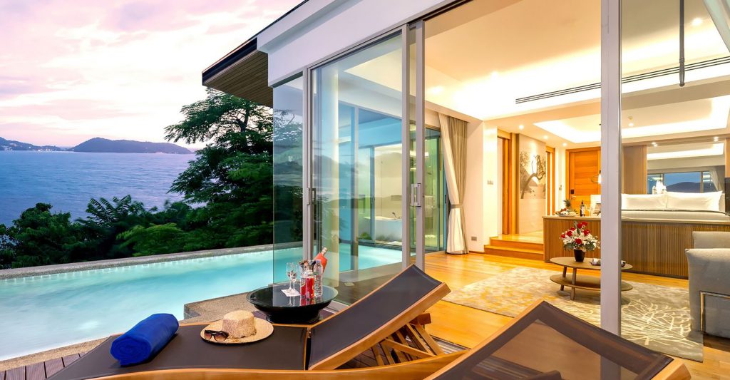 A luxurious poolside suite with lounge chairs, a table with drinks and a sunhat, a private infinity pool, and large glass doors opening to a modern bedroom, overlooking the ocean and lush greenery at sunset.