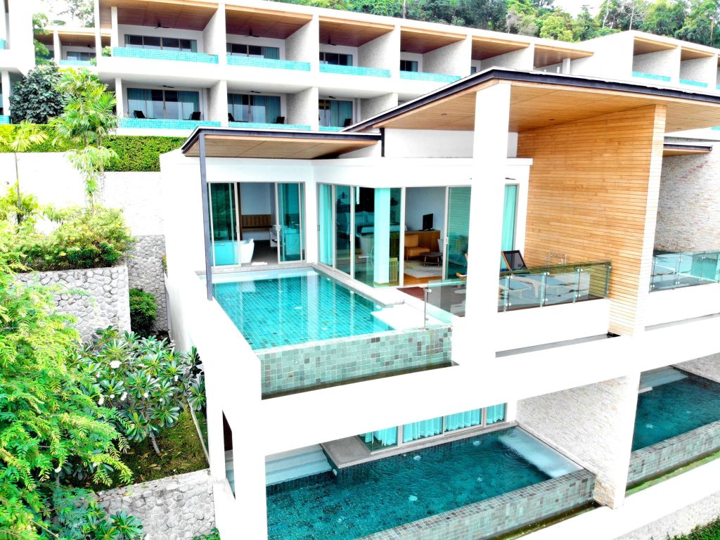 Modern luxury villa with a private infinity pool on a balcony, surrounded by greenery, and part of a multi-level resort complex with glass railings and large windows.